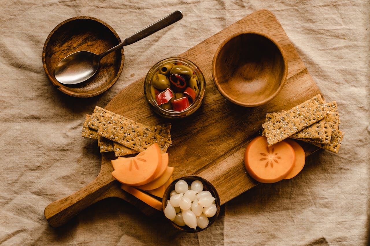 A rustic food setup featuring crackers, olives, and persimmons on a wooden board.